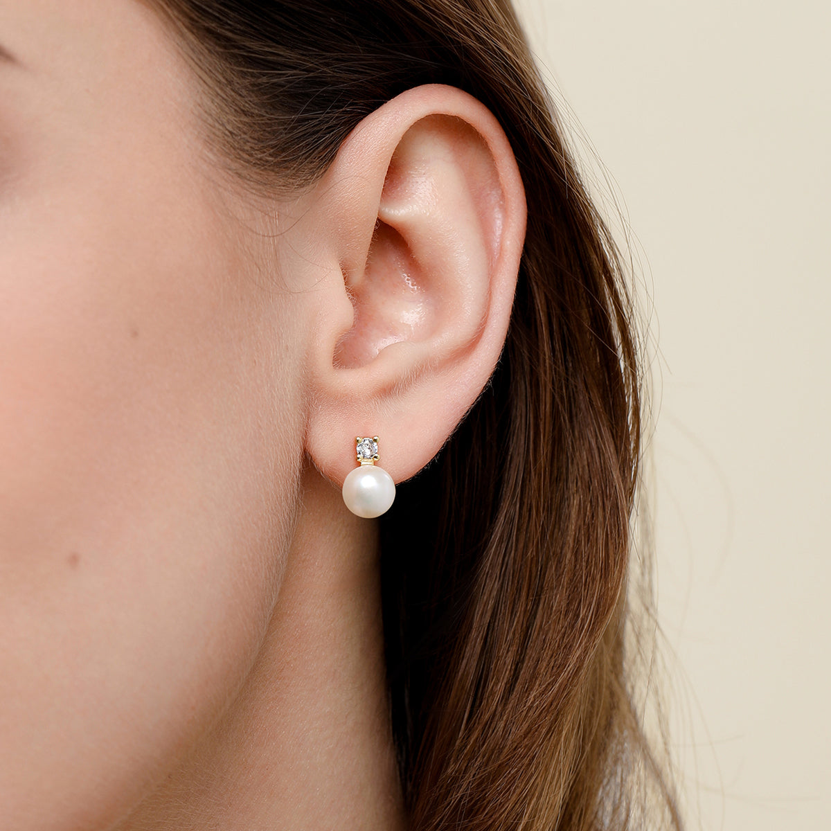 A close-up of a woman's ear wearing a tiny pearl stud earring with a clear rhinestone setting, against a plain light beige background.