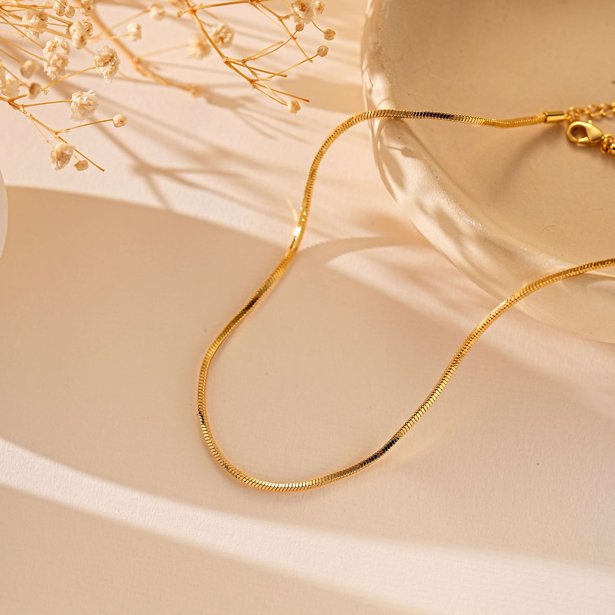 A gold snake chain necklace draped elegantly over a warm beige surface, styled with dried baby's breath flowers and a soft cream dish in the background.