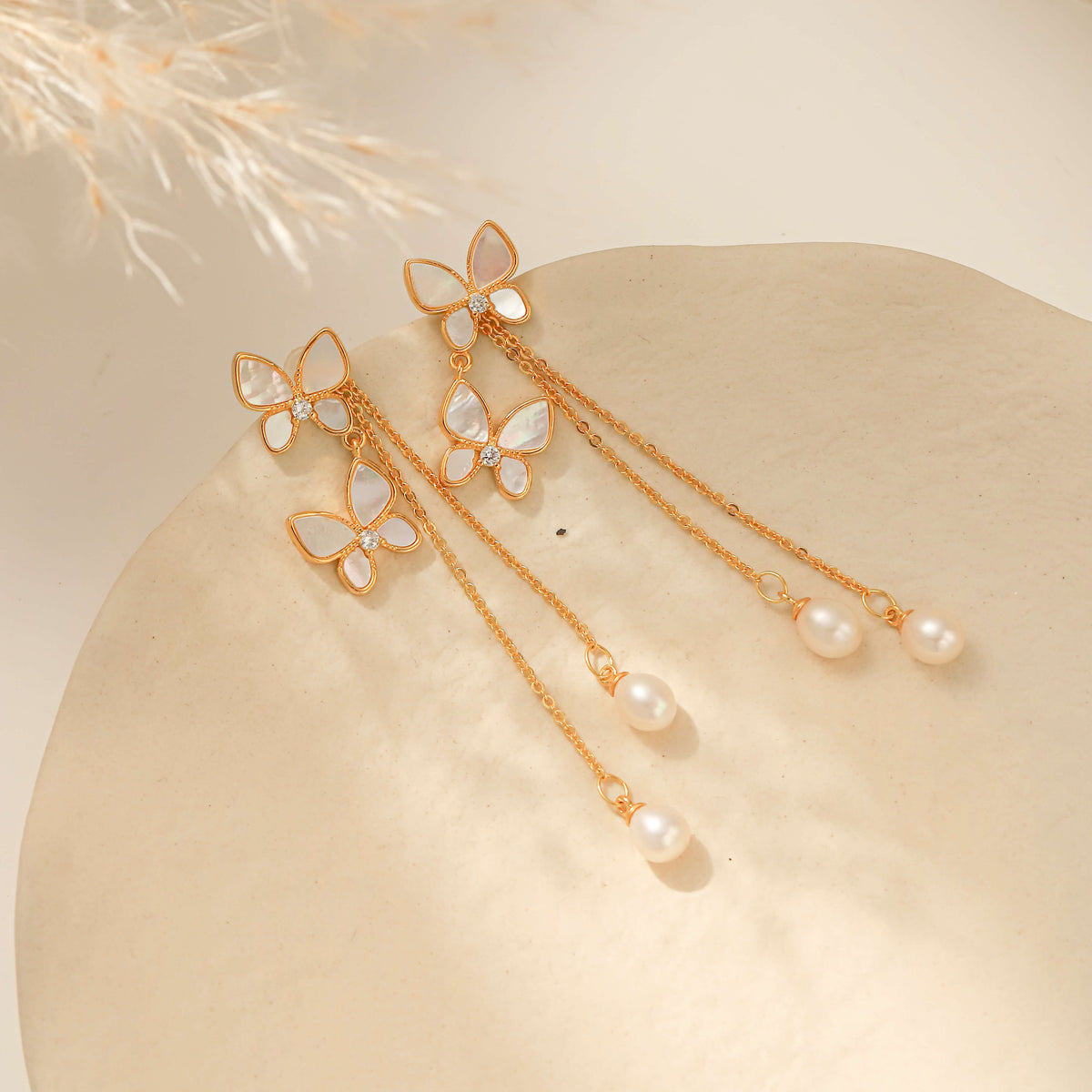 A pair of gold-tone butterfly dangle earrings with white pearl-like accents, displayed on a beige stone surface with dried grass in the corner.