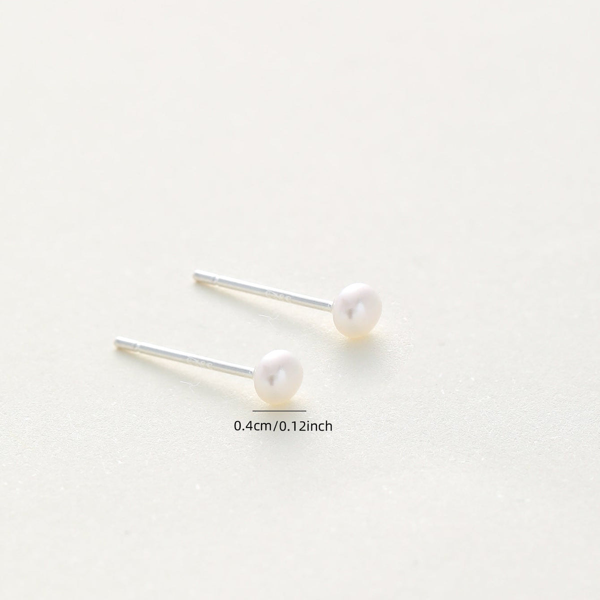 A pair of minimalist stud earrings featuring round white pearls on thin silver posts, displayed on a clean, light off-white background with a small size measurement overlay reading '0.4cm/0.
