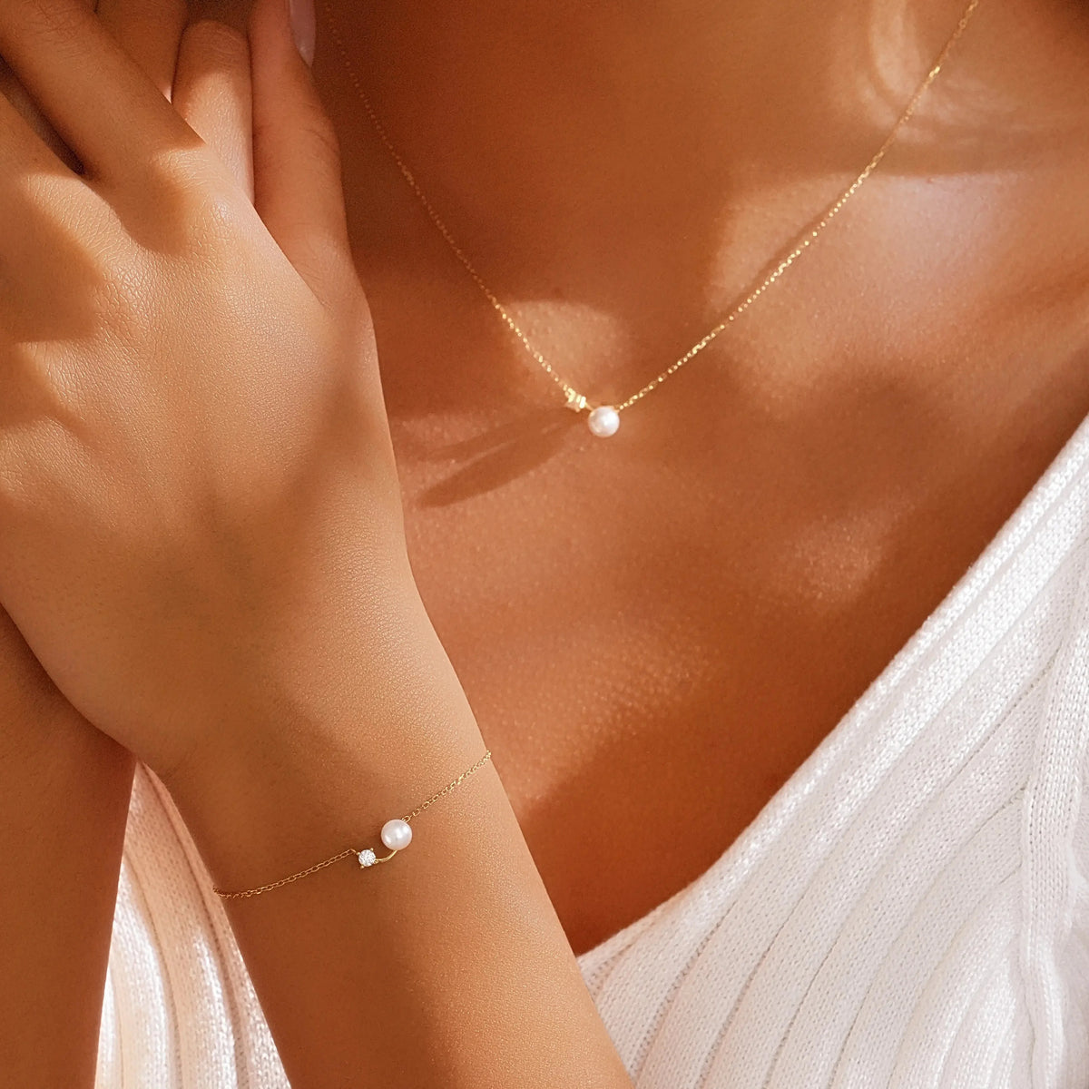 A close-up lifestyle shot of a model wearing a delicate gold necklace and matching gold bracelet, each featuring a small white pearl accent, styled with a white ribbed top.