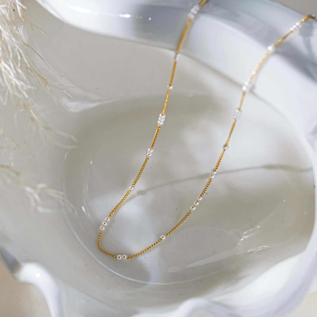 A delicate gold chain necklace with small white pearl-like beads, draped inside a glossy white ceramic bowl.