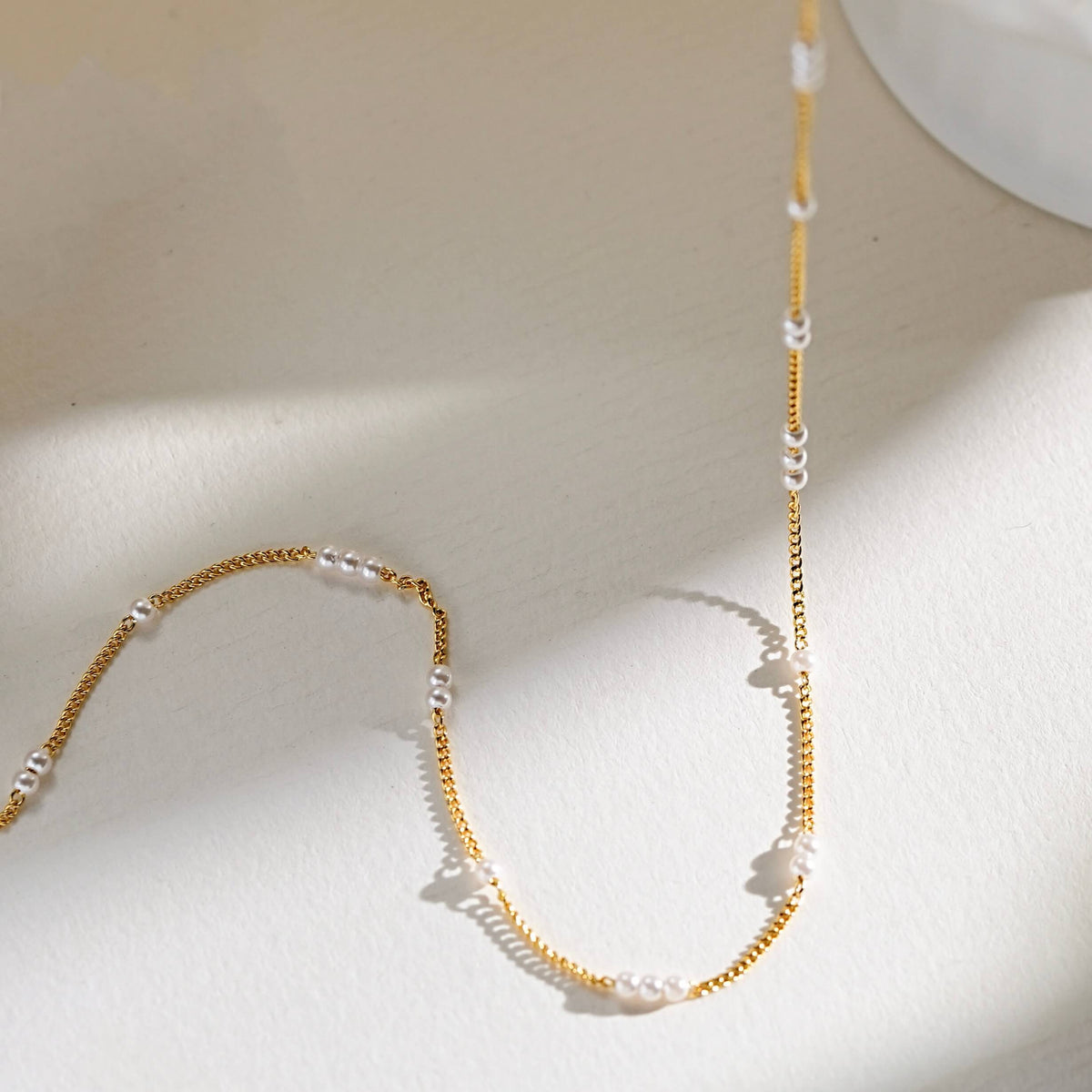 A minimalist gold chain necklace with small white pearl beads spaced along the length, photographed on a warm off-white surface with soft shadows.