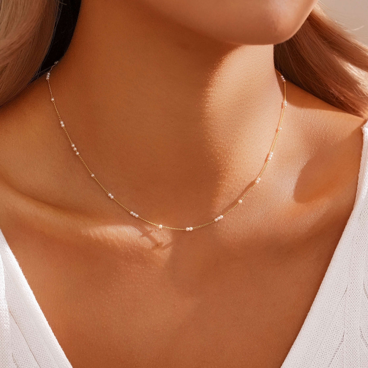 A delicate gold chain necklace with small white bead accents worn on a model's neck, paired with a white top.