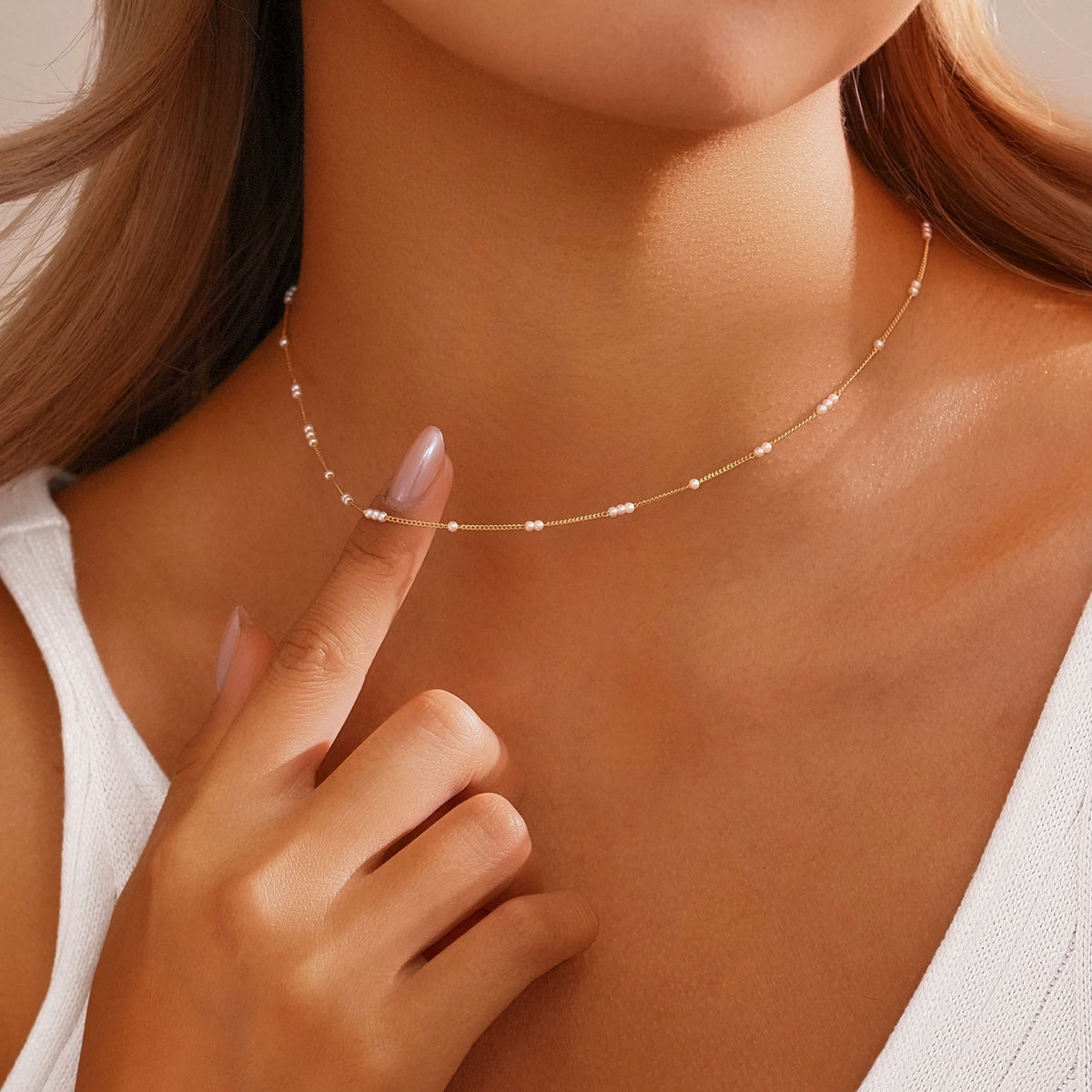 A close-up lifestyle shot of a model wearing a delicate gold chain necklace with small white bead accents, with her finger pointing to a section of the necklace.