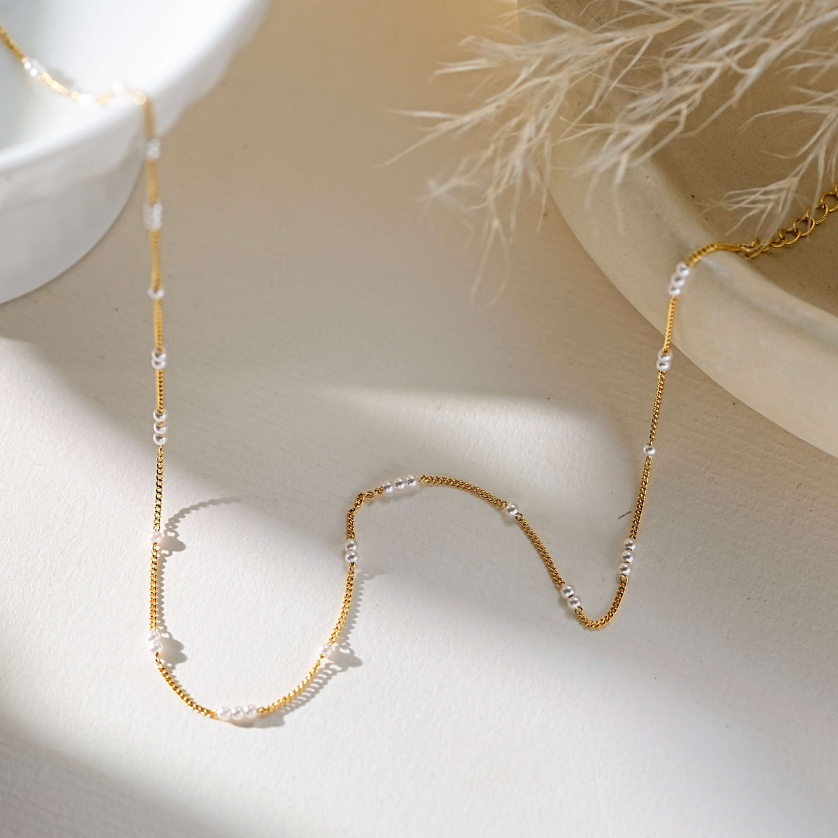 A delicate gold chain necklace with small white pearl-like beads spaced along the chain, styled on a warm cream surface with soft shadows and dried white grass in the background.