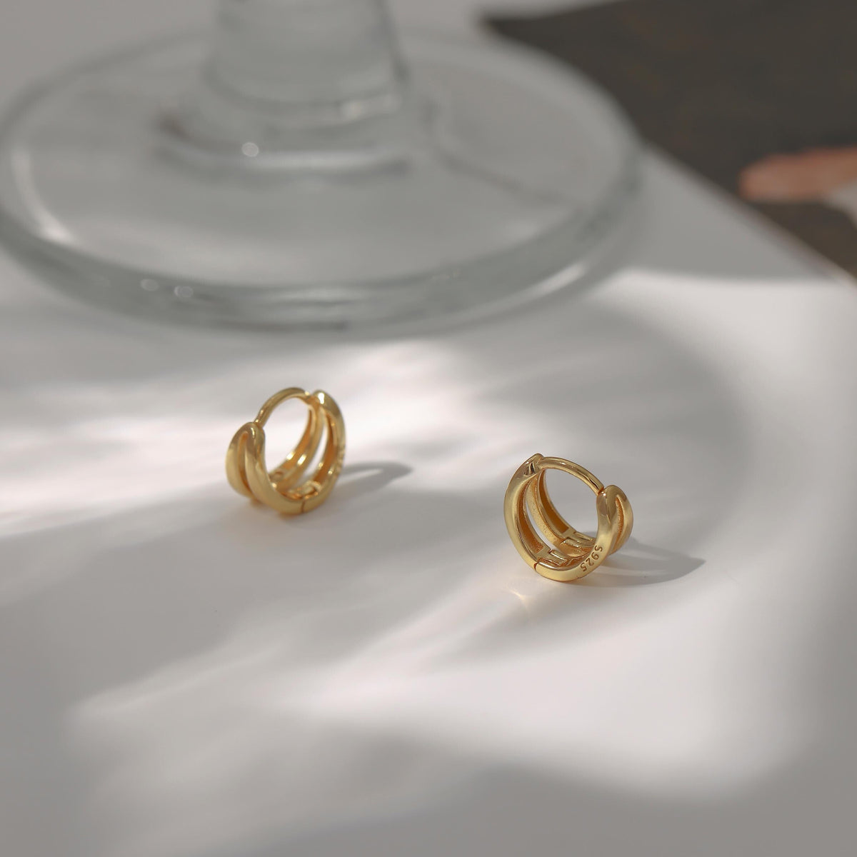 A pair of gold huggie hoop earrings with a textured, ridged finish, displayed on a white surface with artistic shadows and a glass base in the background.