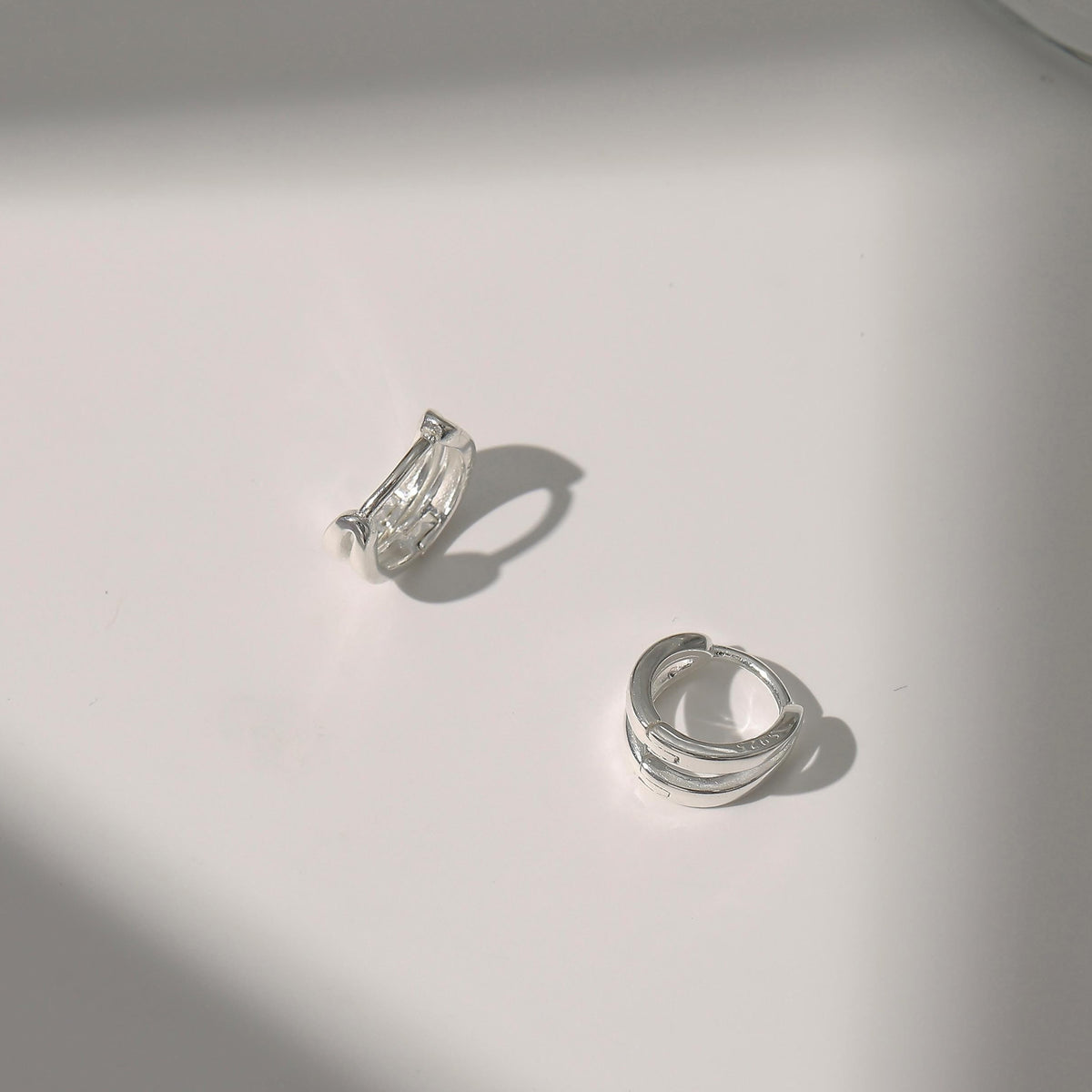 925 solid sterling silver huggie hoop earrings on a white surface with soft shadows. One earring has a textured, hammered finish, and the other is smooth and polished.