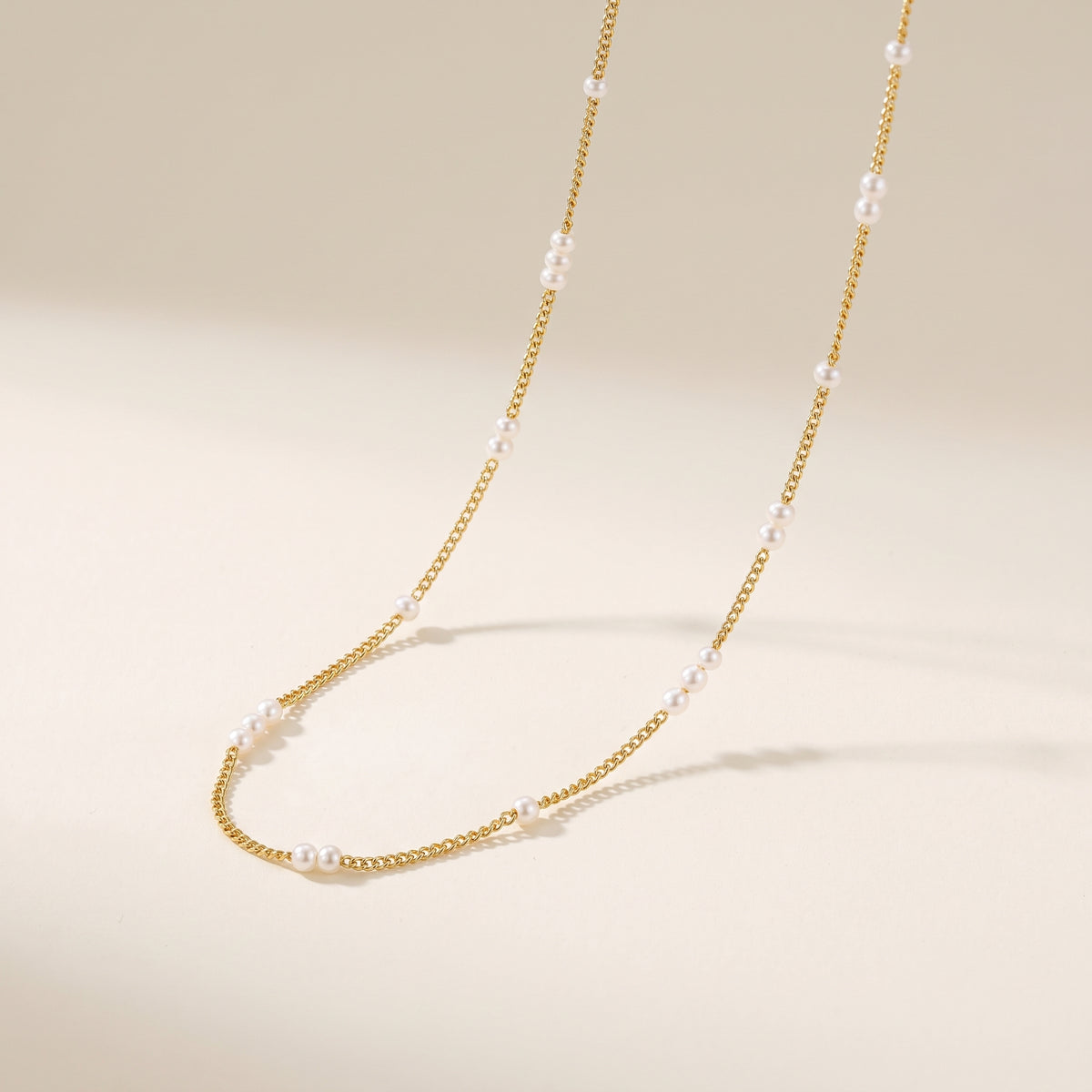 A dainty gold choker necklace adorned with 2mm white pearls, elegantly draped on a warm beige background.