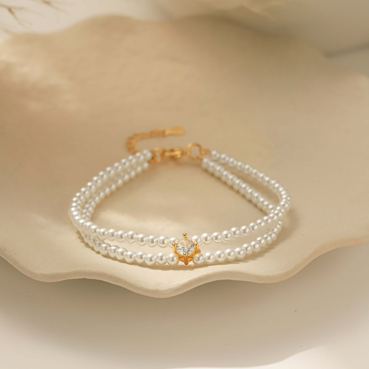 A double layer pearl beaded bracelet with a small gold cz charm, displayed on a scalloped cream ceramic dish.