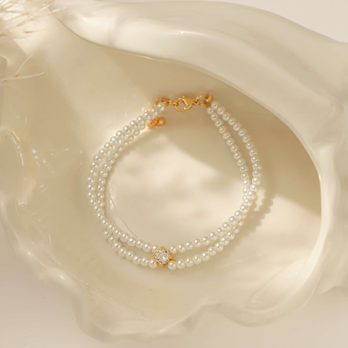 A double-strand pearl bracelet with gold-toned metal details and a central decorative gold clasp, displayed inside a large white clam shell.