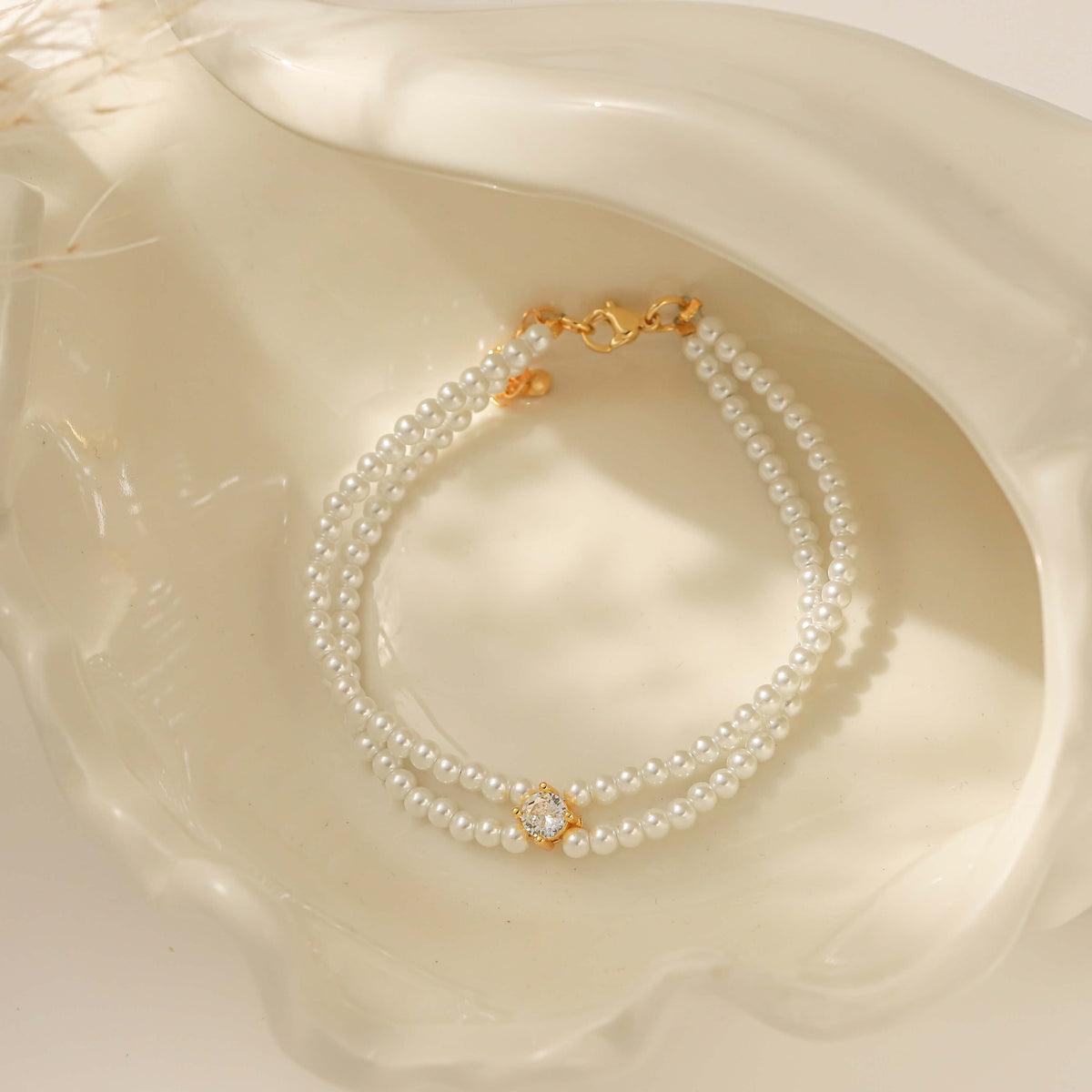 A double-strand pearl beaded bracelet with gold-toned metal details and a central clear stone, displayed inside a white seashell.