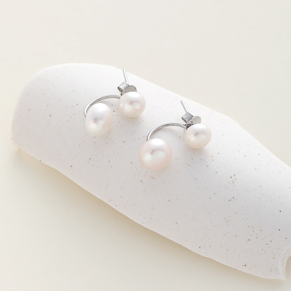 A pair of silver earrings with double white pearls, displayed on a white speckled curved stand against a warm off-white background.