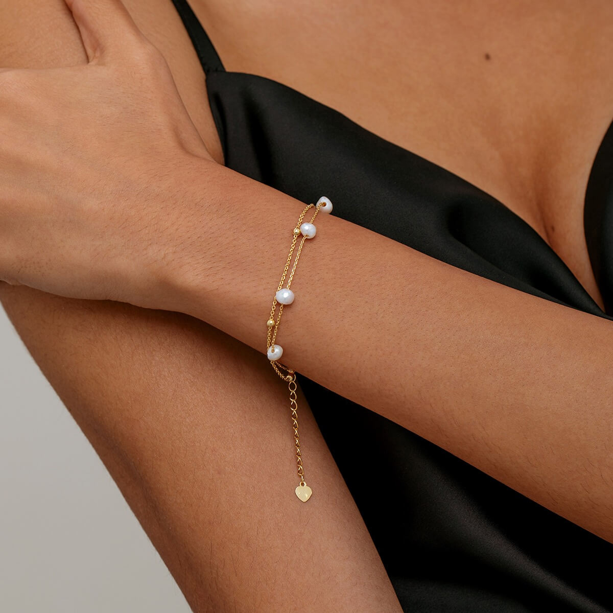A delicate gold double layer chain bracelet with small white pearls and a dangling heart charm, shown worn on a model's wrist.
