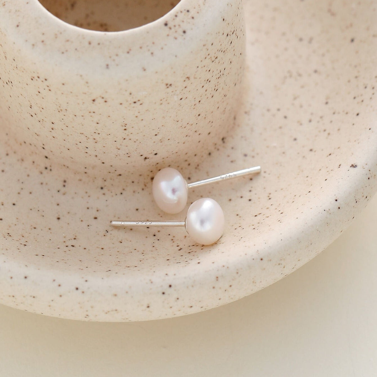 A pair of minimalist stud earrings with lustrous white freshwater pearls on silver posts, displayed on a speckled beige ceramic dish.