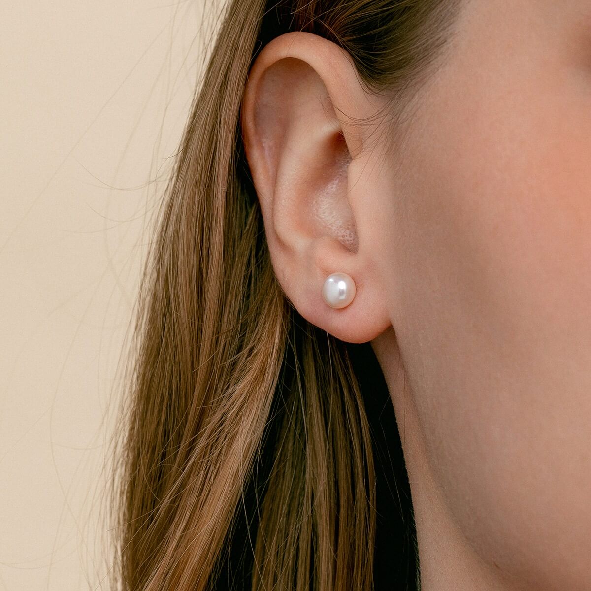A close-up of a model's ear wearing a minimalist white pearl stud earring against a soft beige background.