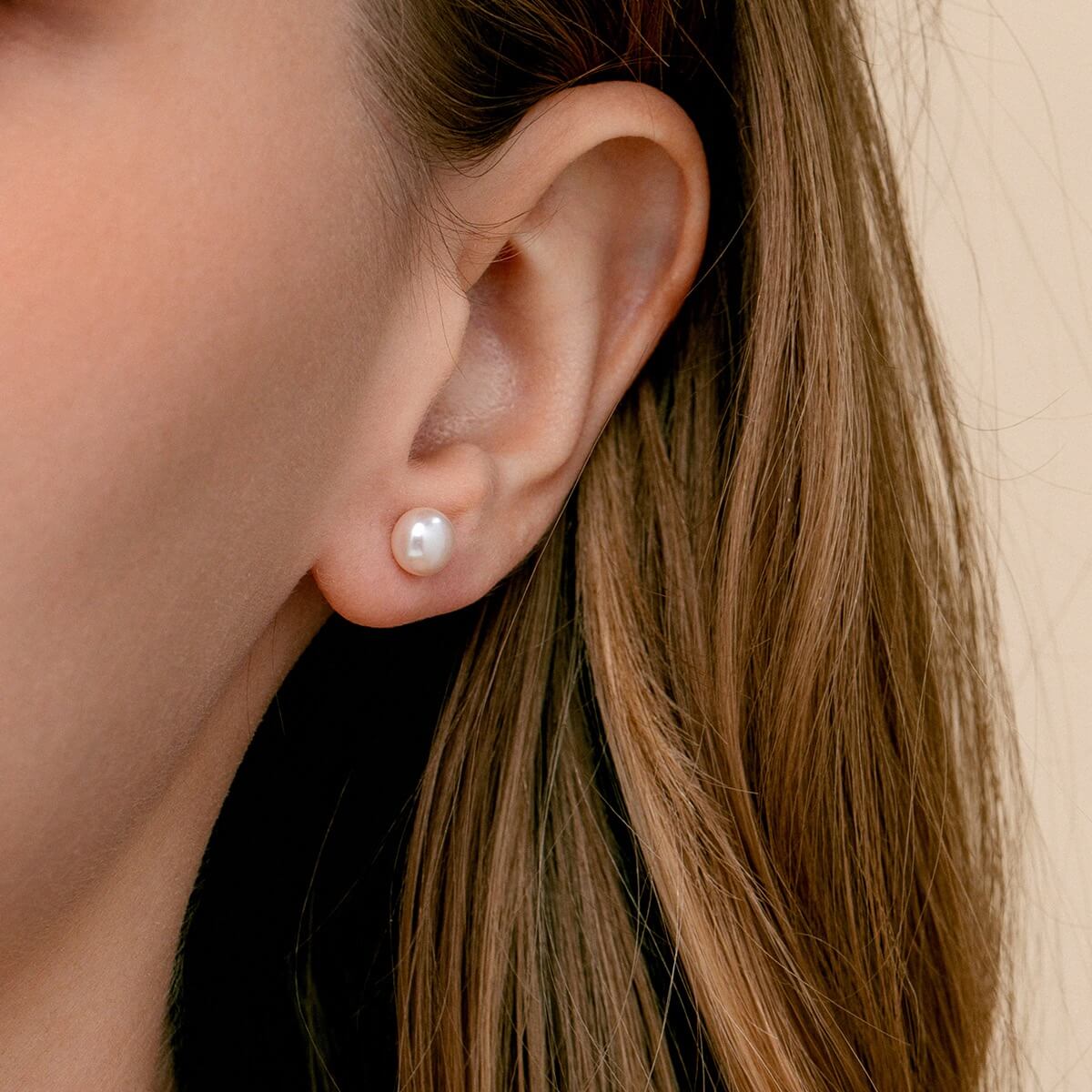 A close-up of a woman's ear wearing a single round white pearl stud earring against a soft beige background.