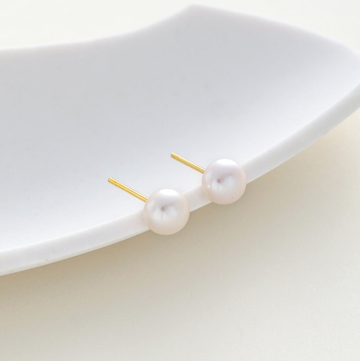 A pair of dainty gold stud earrings with round white freshwater pearls, displayed on a white ceramic dish against a light cream background.