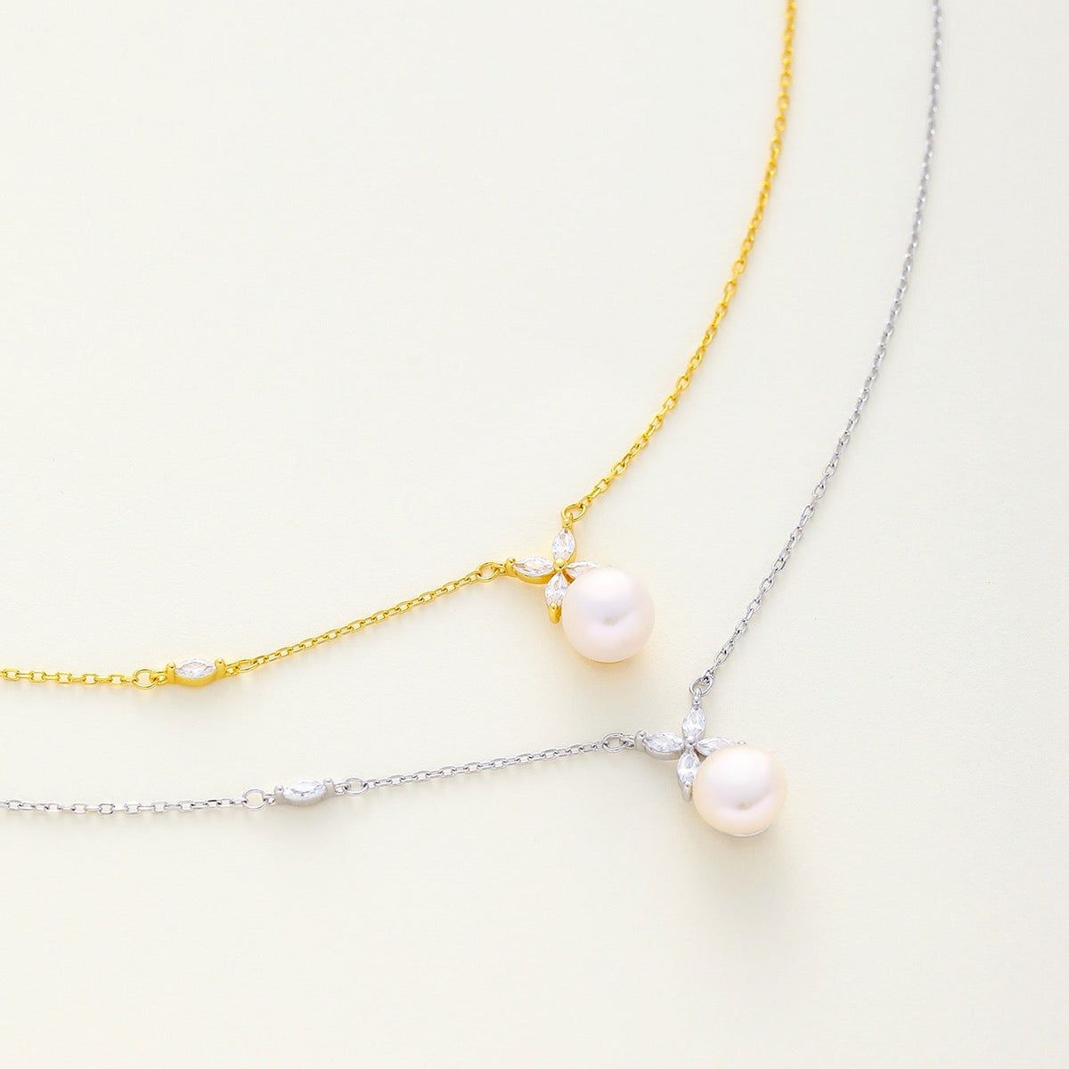 Two dainty pearl and floral cz necklace laid on a light cream background, available in gold and silver finish
