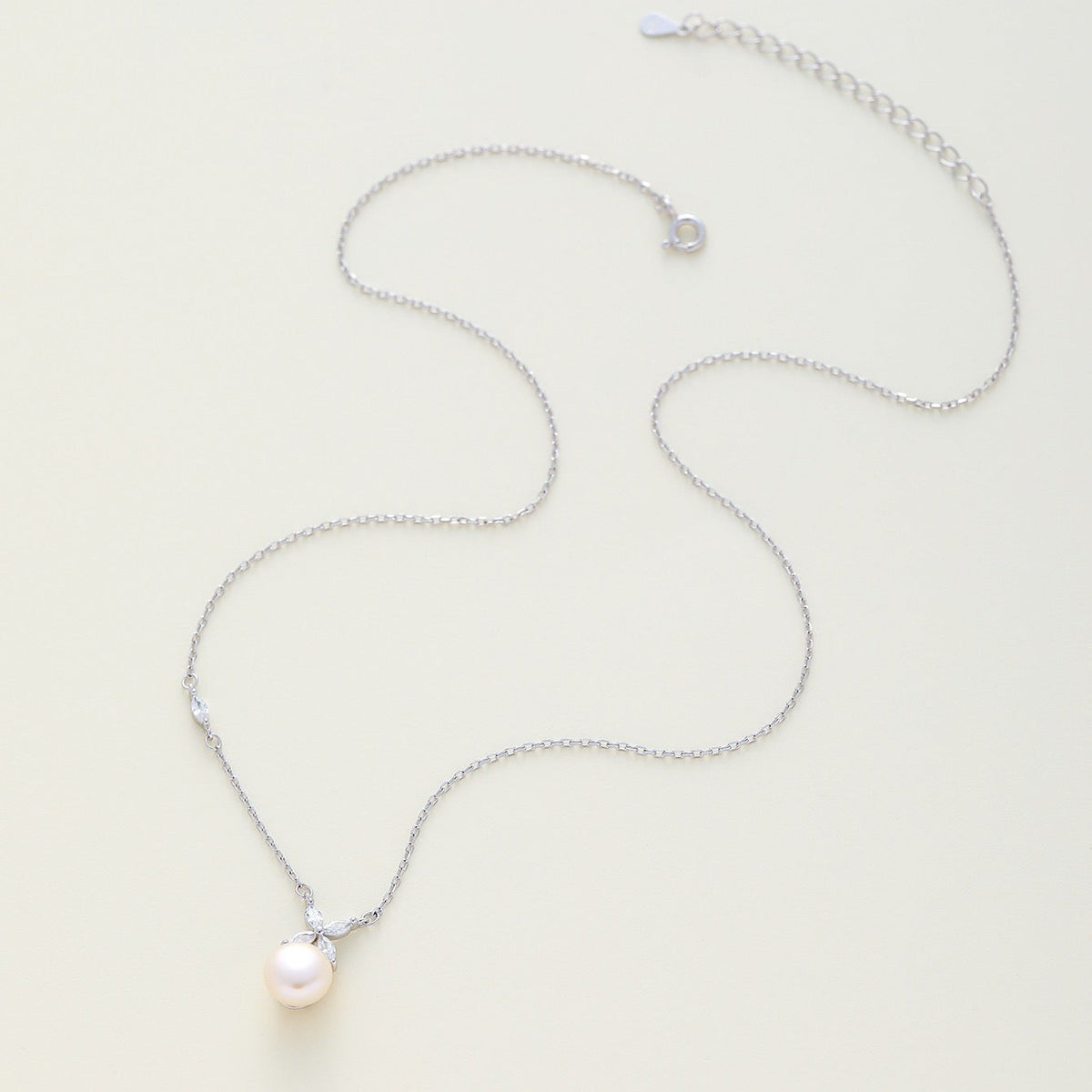 A delicate silver chain necklace featuring a single white freshwater pearl pendant, displayed on a light cream background.