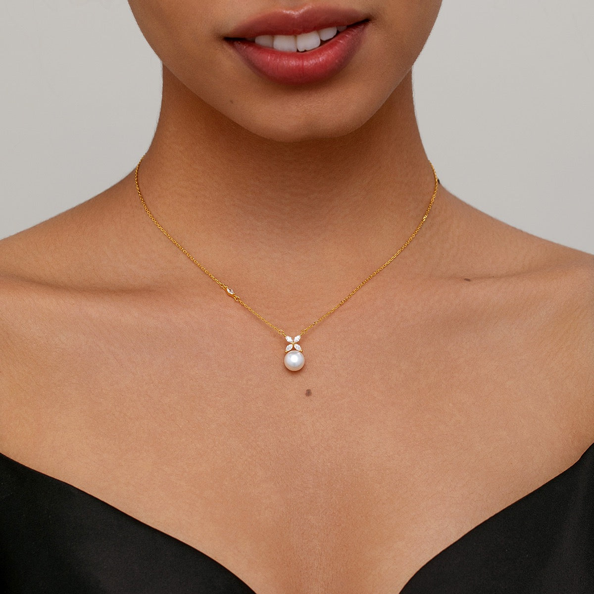 A close-up of a model wearing a delicate gold chain necklace with a small flower charm and a single white pearl pendant, styled with a black top against a neutral gray background.