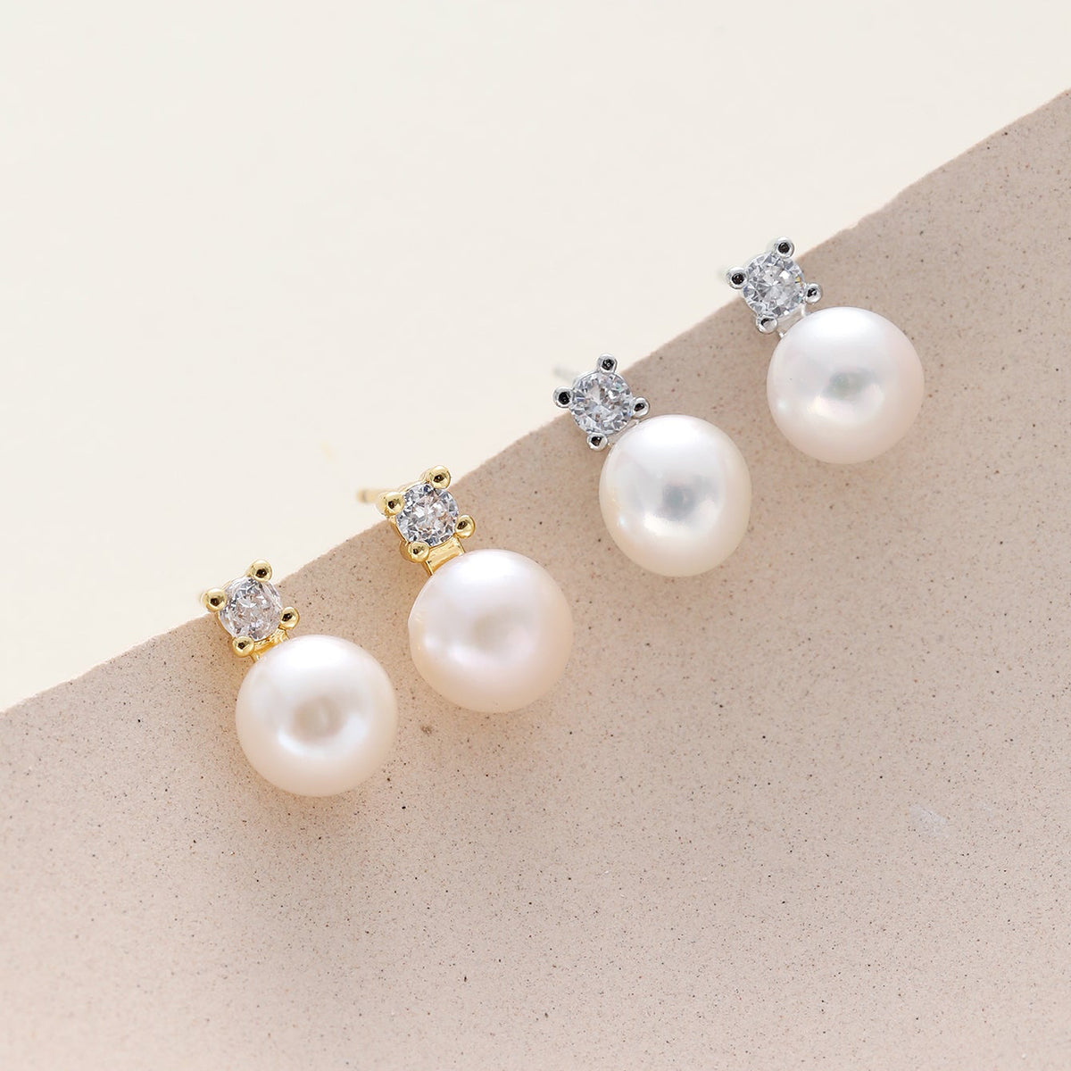 Dainty cz pearl stud earrings displayed on a beige surface, featuring large white pearls suspended from small clear stones in gold and silver settings.