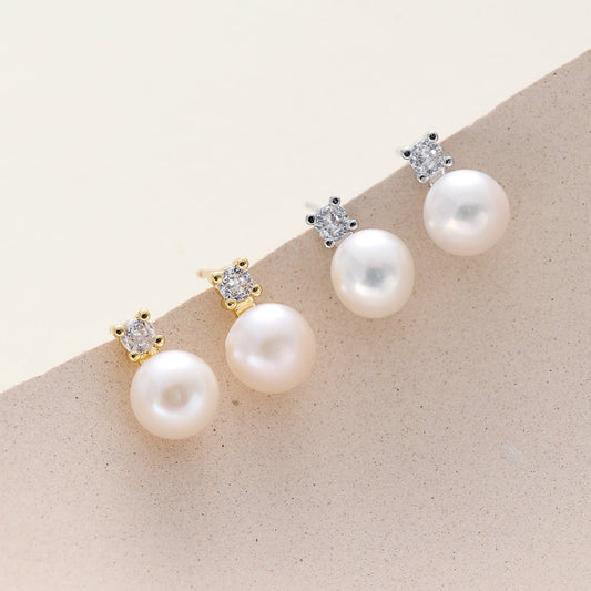 Dainty cz pearl stud earrings displayed on a beige surface, featuring large white pearls suspended from small clear stones in gold and silver settings.