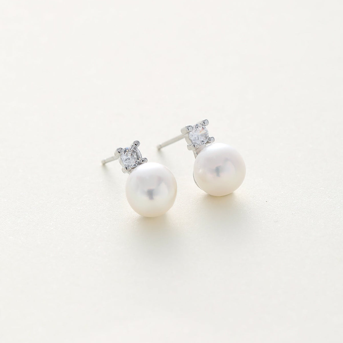 A pair of silver stud earrings featuring a small clear gemstone in a square setting above a round white pearl, shown on a clean off-white background.