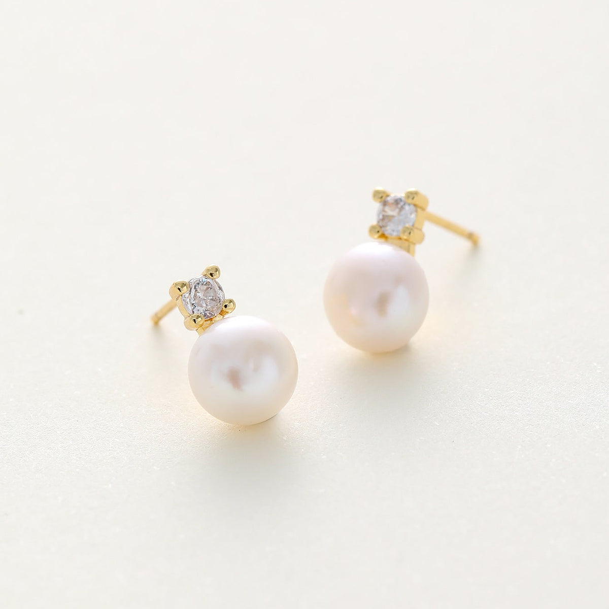 A pair of gold stud earrings featuring a small cz in a star-like setting above a round white pearl, shown on a light cream background.