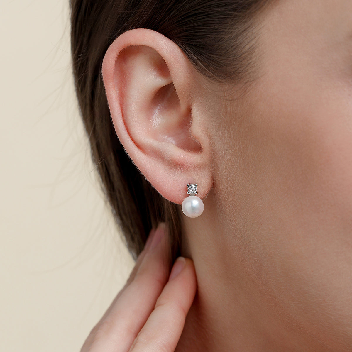 A close-up of a woman's ear wearing a dainty minimalist silver stud earring with a white pearl and a clear rhinestone setting, against a soft beige background.