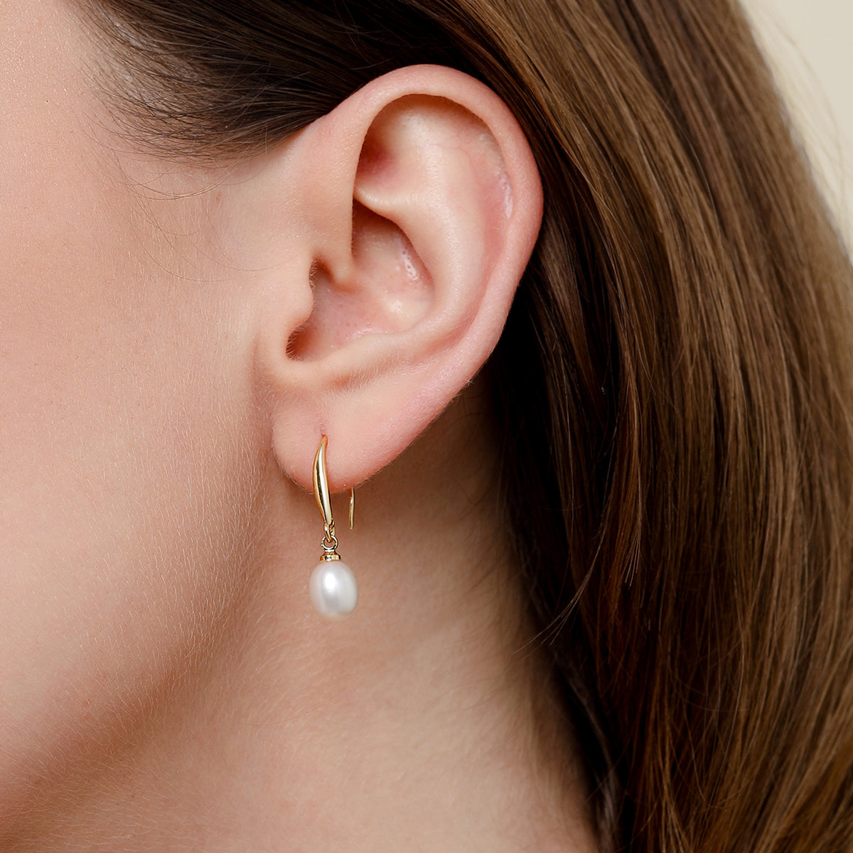 A close-up of a model's ear wearing a delicate gold hook earring with a single dangling white freshwater pearl.