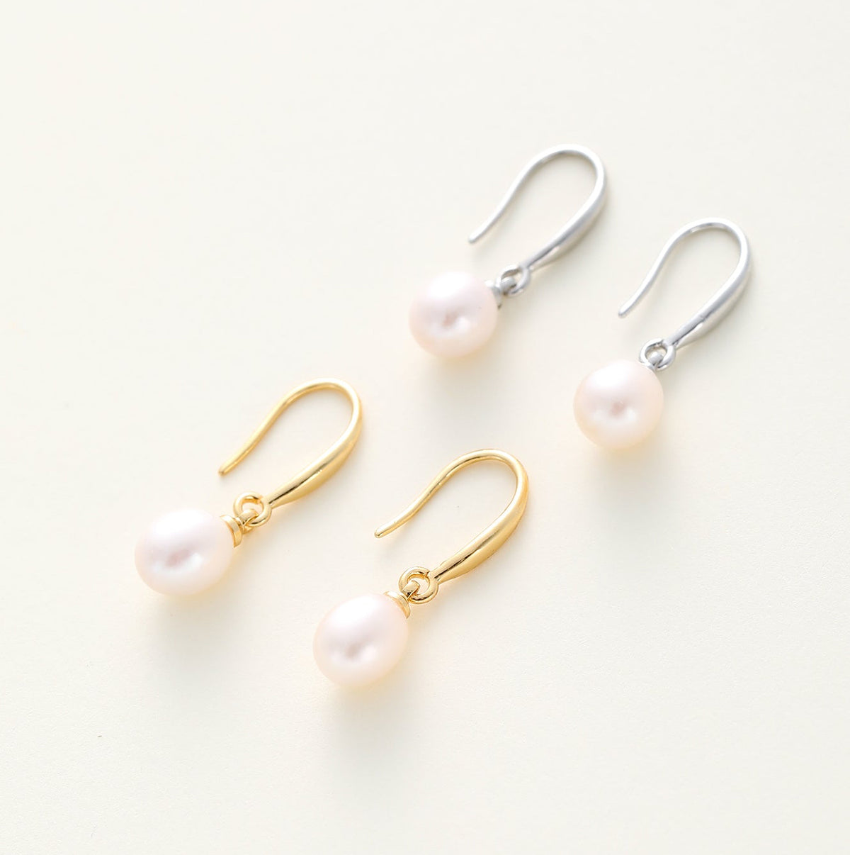  drop earrings with round white pearls and curved metal hooks, shown in gold-tone and silver-tone finishes on a light cream background.