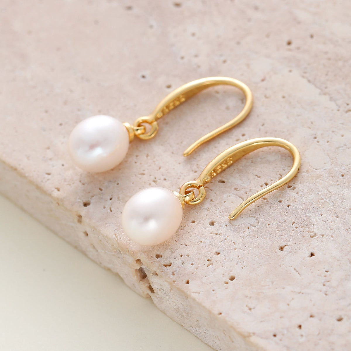 A pair of gold drop earrings with round white freshwater pearls, displayed on a textured beige stone surface.