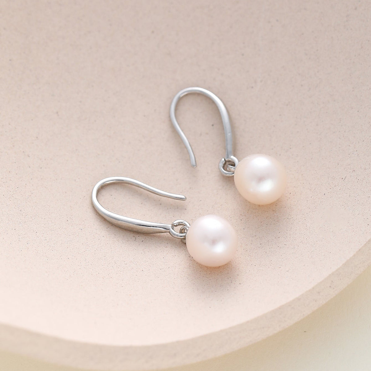 A pair of dainty silver hook dangle earrings with round, lustrous white freshwater pearls, displayed on a light beige textured surface.