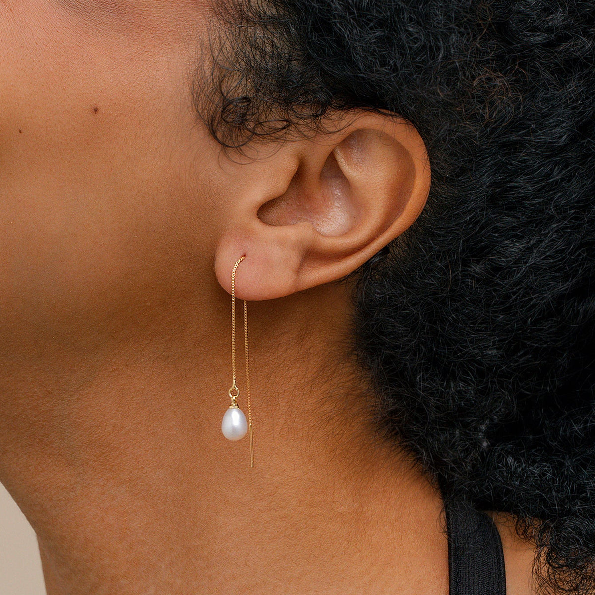 A close-up of a women’s ear wearing a gold threader drop earring with a teardrop-shaped white pearl.