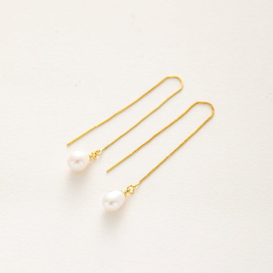 A pair of gold dangle earrings with a single small white pearl drop, photographed on a clean, off-white background.
