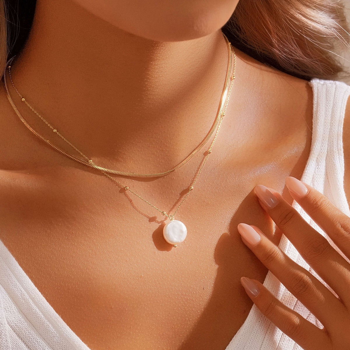 A close-up of a woman wearing a delicate double-layered gold necklace with a round white pearl pendant, styled with a white sleeveless top under warm sunlight.