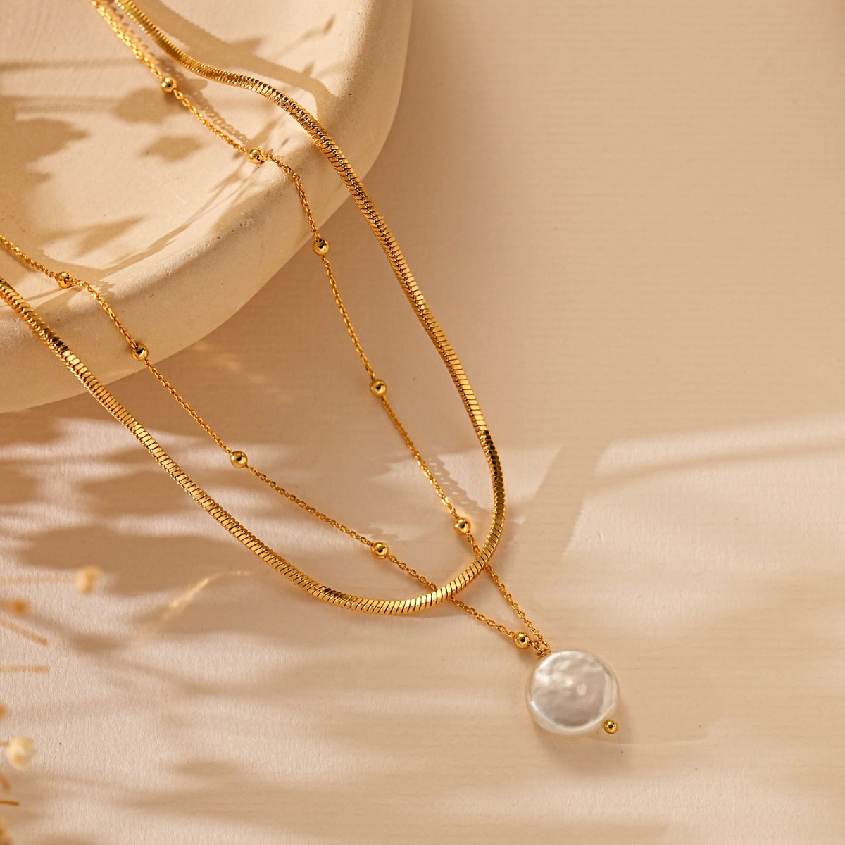 A double-layered gold necklace with a textured snake chain and a delicate chain with small beads, featuring a round white pearl pendant, styled on a beige fabric background with soft shadows.