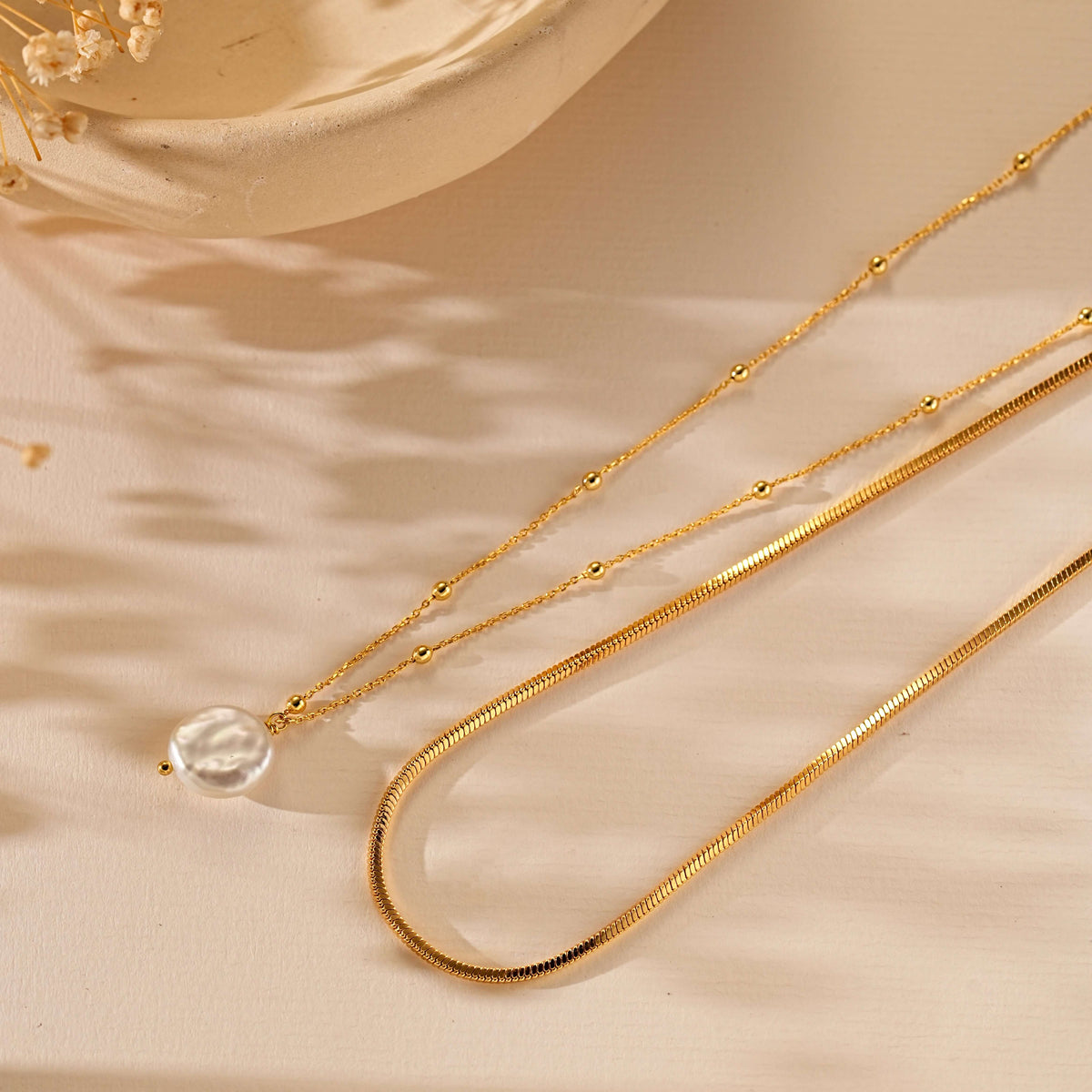 A delicate gold necklace with a small clear faceted bead pendant, displayed on a warm beige fabric background with soft shadows.