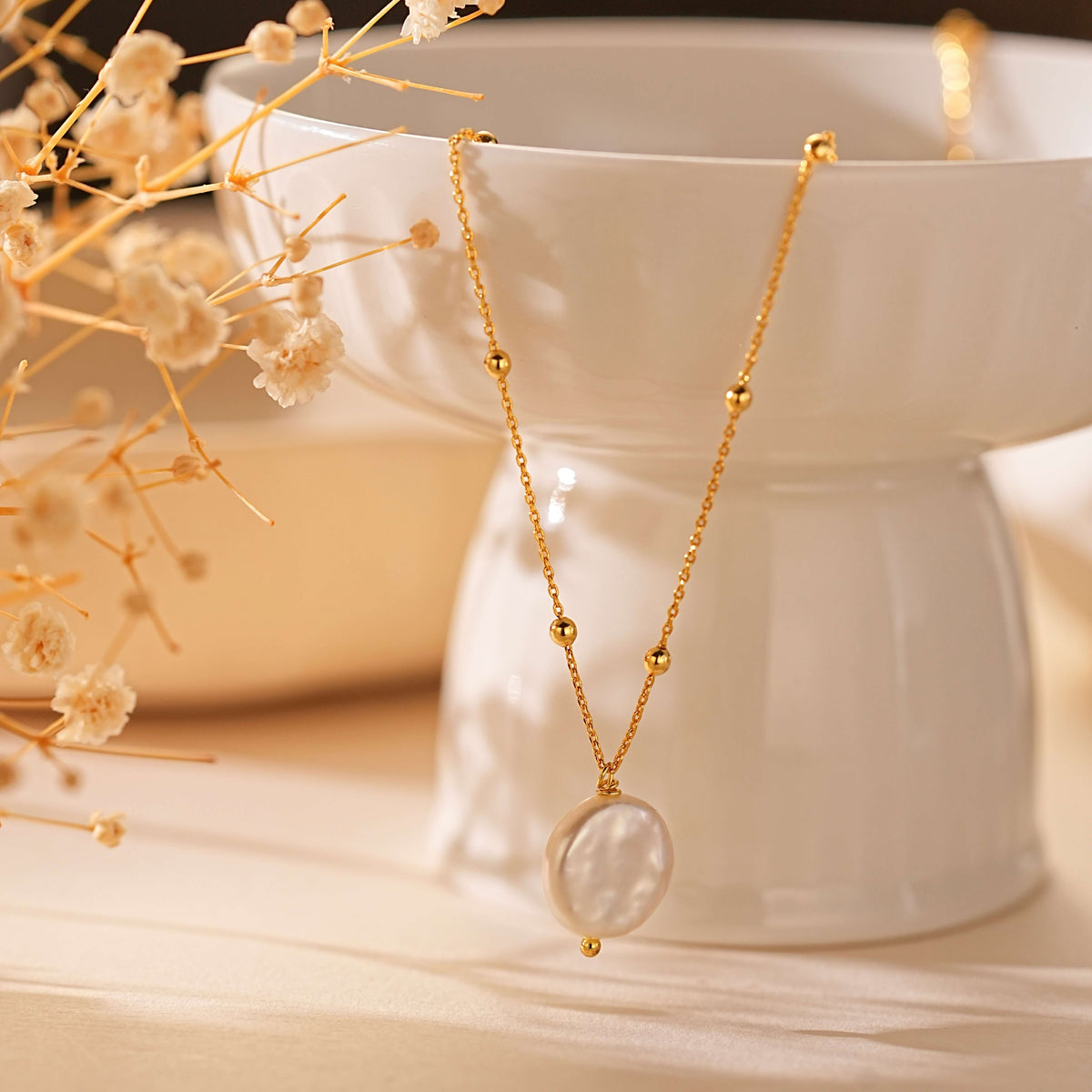 A gold chain necklace with a large oval mother-of-pearl pendant, draped over a white ceramic pedestal with dried baby's breath flowers in the foreground.