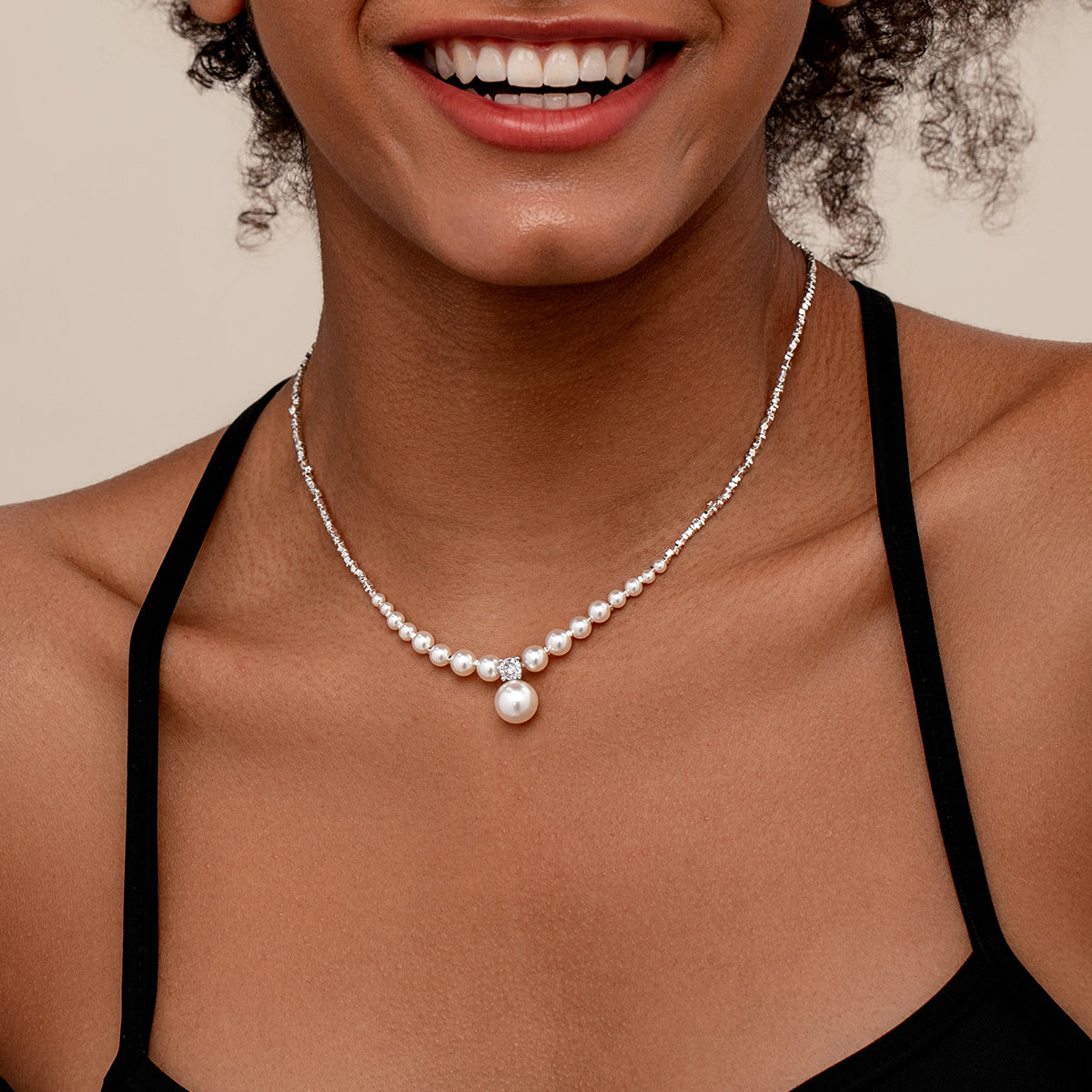 A close-up of a model wearing a delicate silver beaded necklace with a central cluster of white pearls and a larger drop pearl pendant, styled with a black halter top against a warm beige background.