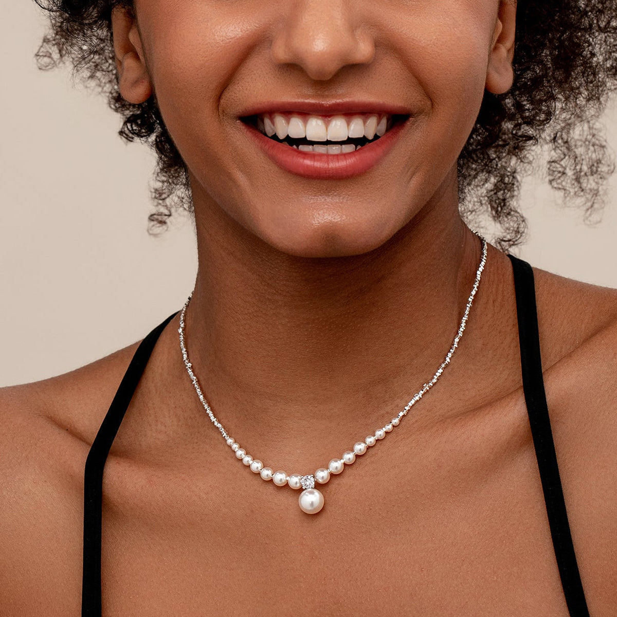 A smiling woman with curly hair wearing a bridal silver necklace with a row of white pearls and a single larger pearl pendant with CZ, shown against a plain beige background.