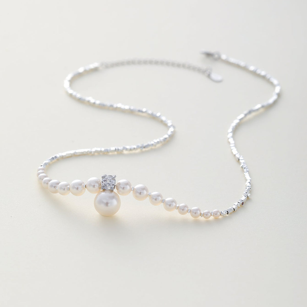 A delicate silver chain necklace featuring a strand of white lab pearls, a central cluster of small pearls and a clear crystal, and a larger pearl pendant, displayed on a light cream background.