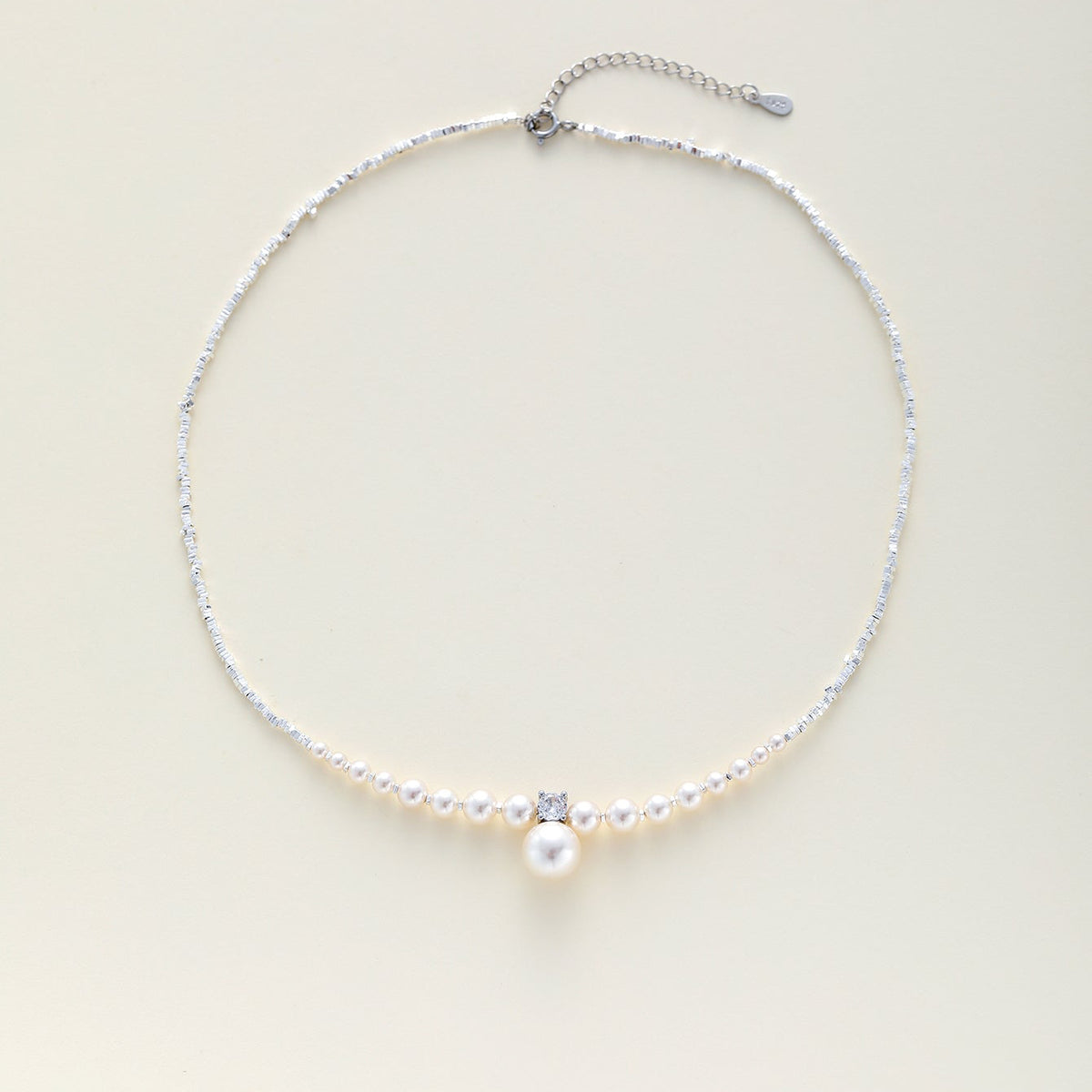 A delicate silver chain necklace with a row of small white pearls, a central clear rhinestone, and a larger white pearl pendant, displayed on a light cream background.