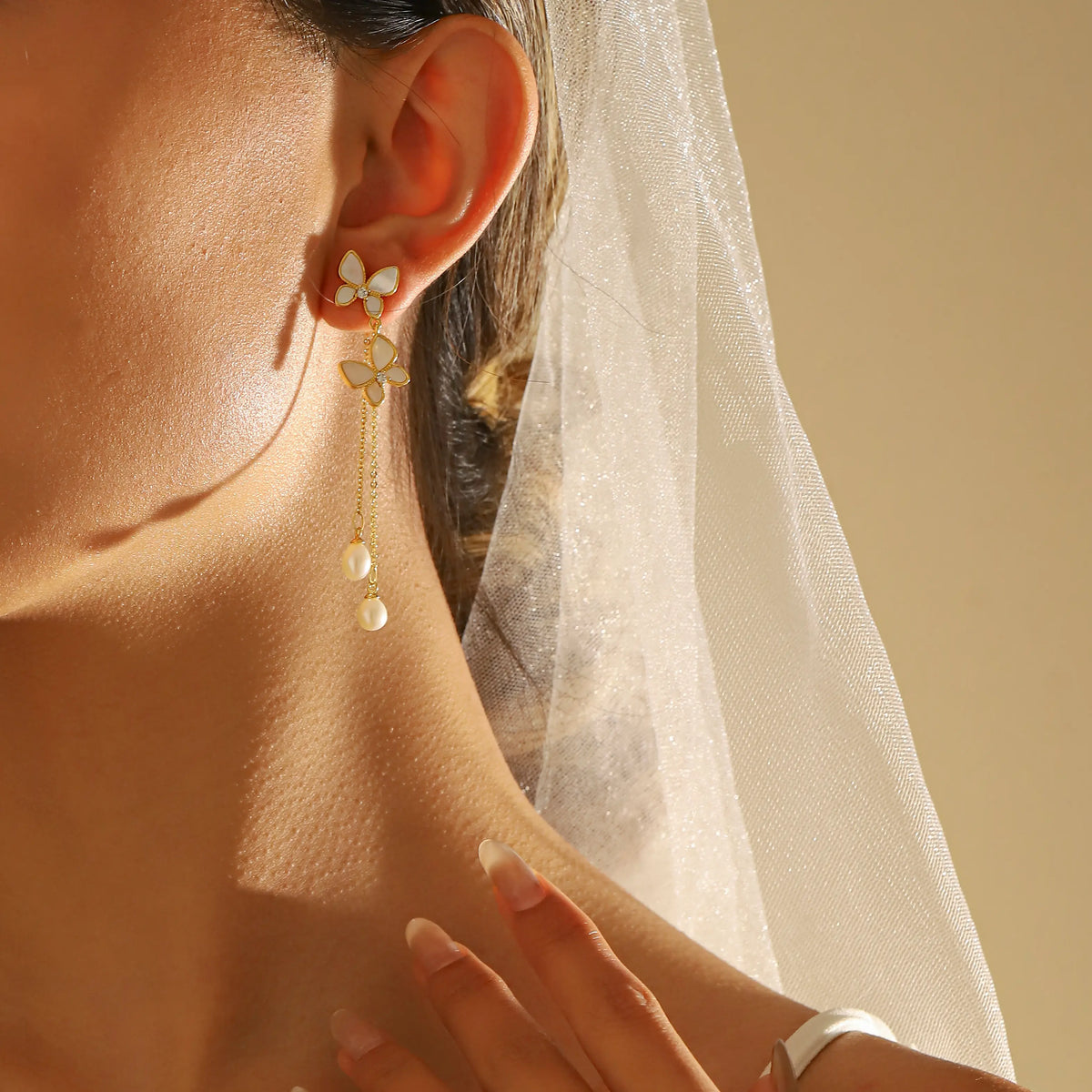A close-up of a woman wearing a delicate gold dangle earring with white enamel butterfly details and small pearl-like drops, styled with a white bridal veil under warm sunlight.