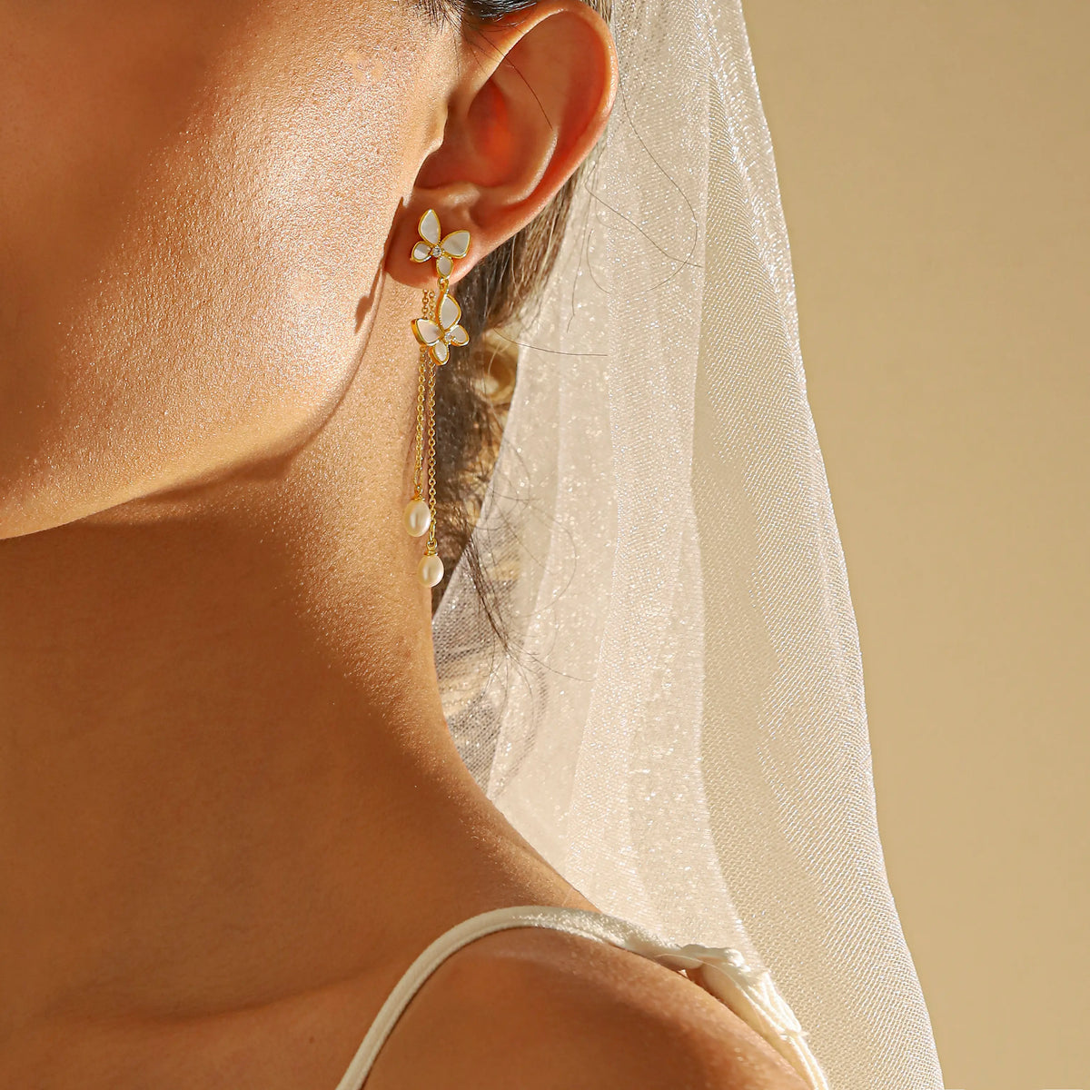 Close-up of a bride's ear wearing a gold floral drop earring with white pearl-like accents, with a white bridal veil and bare shoulder visible against a warm beige background.