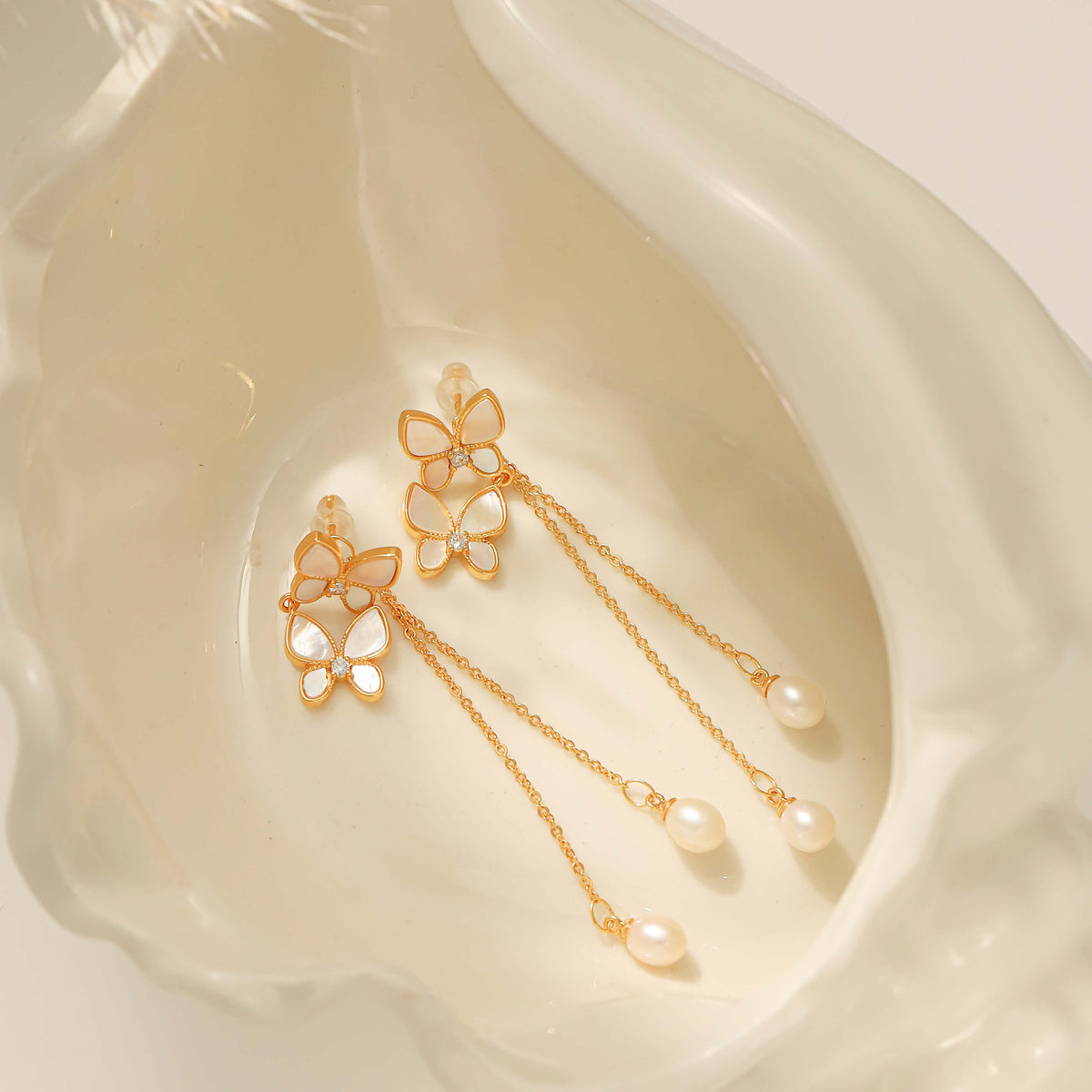 A pair of gold butterfly dangle earrings with white pearl-like drops, displayed inside a white conch shell.