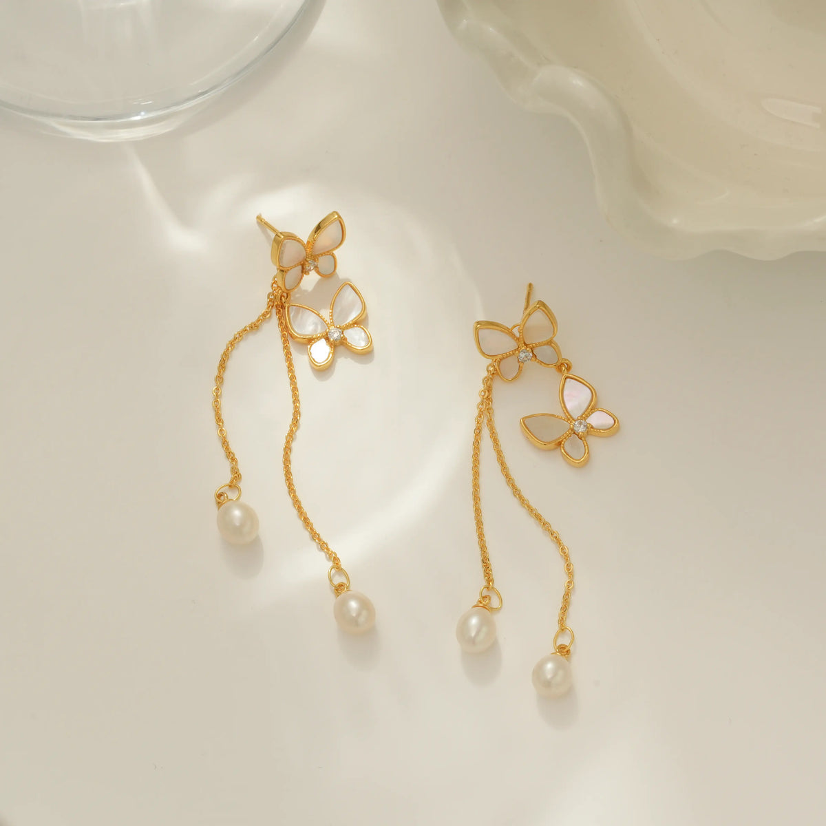 A pair of gold dangle earrings with butterfly-shaped charms, white pearl inlays, and hanging pearl beads, displayed on a clean white surface with soft shadows.