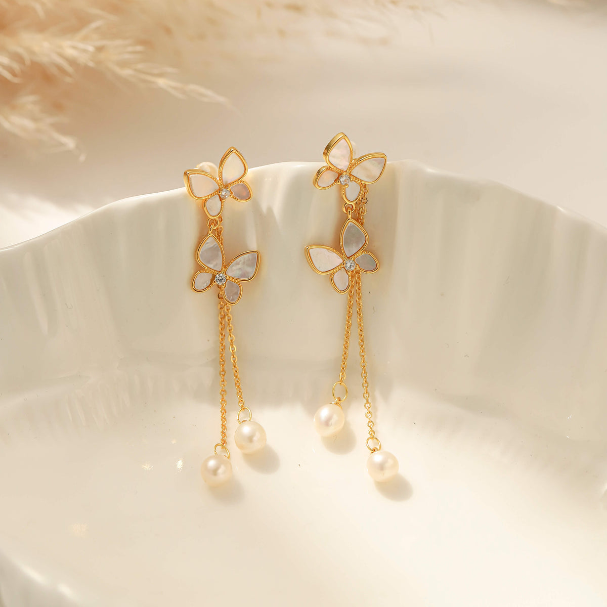 A pair of gold-tone dangle earrings with mother-of-pearl butterfly motifs and hanging pearl beads, displayed on a white ceramic dish.