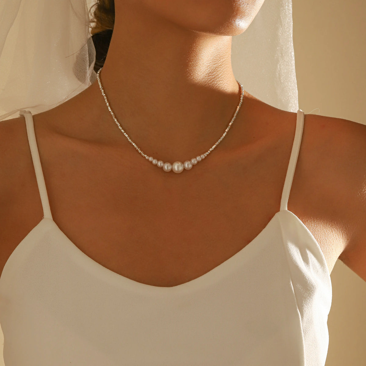 A close-up of a woman wearing a delicate pearl necklace with three larger central pearls, styled with a white spaghetti-strap camisole and a sheer white veil.